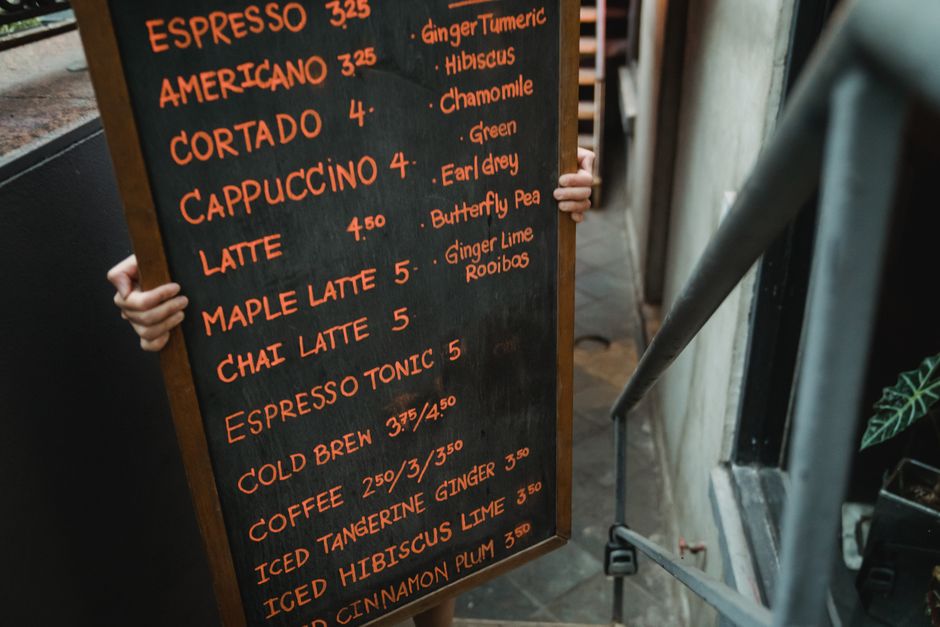 A person holding a large black chalkboard menu outside a cafe or restaurant, displaying drink options written in orange chalk, including espresso, americano, cortado, cappuccino, and latte, along with various flavoured teas such as ginger turmeric, hibiscus, chamomile, and green tea. The menu lists prices next to each item. The person's hands are visible gripping the edges of the board, which is positioned against a building wall on a sidewalk, with a metallic handrail and potted plant in the foreground. The setting appears to be an urban street environment, possibly near a house or small commercial property. The image is associated with local coffee shop activities but also relates to the broader context of home or small business relocations, as part of transport or packing processes handled by companies like Earls Court Removals.