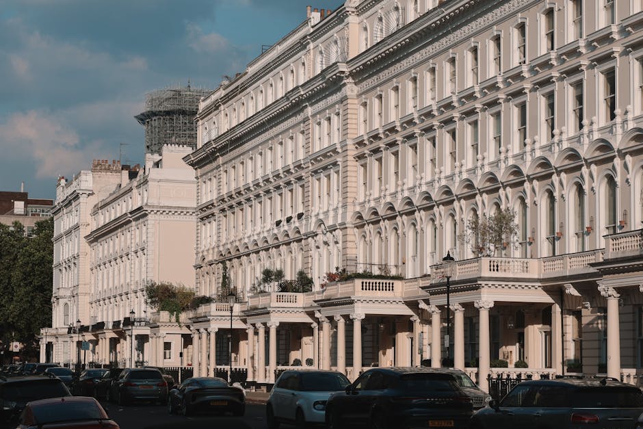 A street view showing a row of white Victorian-style terraced buildings with ornate architectural details, including arched windows, decorative cornices, and balconies with balustrades. In the foreground, multiple cars are parked along the pavement, with some partially visible. The image is taken during daylight under a partly cloudy sky, with a building covered in scaffolding and black netting visible in the background. The scene captures a typical residential area near Earls Court station, with no moving activity visible but representing a setting where house removals, such as packing and furniture transport by Earls Court Removals, would take place during a home relocation process.