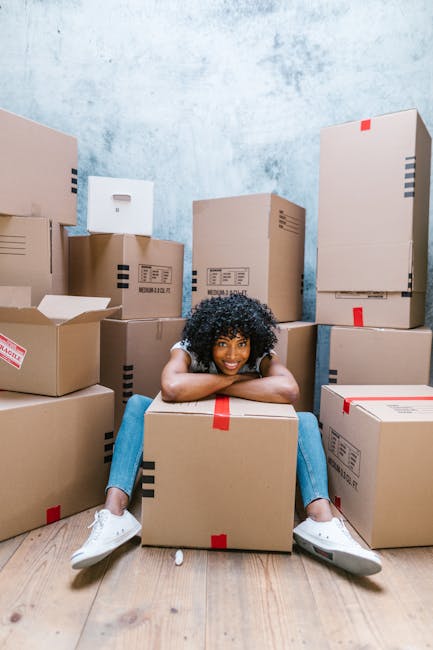 A young woman with curly black hair, wearing a white top, light blue jeans, and white sneakers, is sitting on the floor amidst a large collection of packed cardboard boxes of various sizes in a room with a textured light blue wall and wooden flooring. She is smiling and leaning forward on a medium-sized cardboard box with red packing tape, surrounded by additional boxes stacked against the wall and on the floor, some partially open or sealed with tape. The boxes are labeled with barcodes and shipping information, indicating they are prepared for a home relocation or furniture transport process. The environment suggests a professional removals setting, with the boxes organized for an upcoming move, reflecting a typical packing and moving scene that Earls Court Removals specializes in.