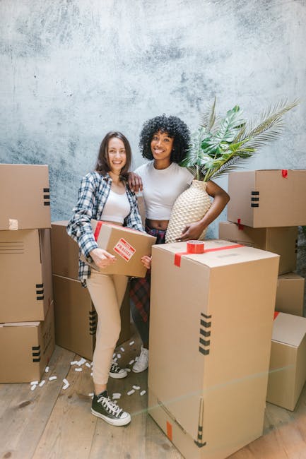 Two women participating in a home relocation process are standing indoors among several packed cardboard boxes, some sealed with red and black tape. The woman on the left, wearing a plaid shirt, beige pants, and black and white sneakers, holds a medium-sized box and smiles at the camera. The woman on the right, dressed in a white t-shirt and patterned trousers, is holding a tall woven vase with a large artificial palm leaf arrangement. The boxes vary in size and are positioned on a light wooden floor against a textured grey wall. The scene depicts the loading or packing phase of furniture transport and packing for a move, with additional boxes stacked behind and around them, highlighting an organized packing and moving environment typical of a professional removals service. Earls Court Removals, a company specialising in house removals, is experienced in managing all aspects of furniture transport and building access for efficient home relocation.