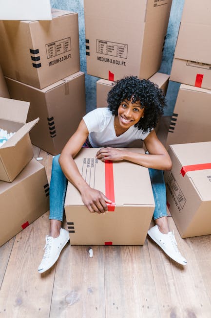 A young woman with curly black hair, wearing a white top, light blue jeans, and white sneakers, is sitting on the floor amidst a large collection of packed cardboard boxes of various sizes in a room with a textured light blue wall and wooden flooring. She is smiling and leaning forward on a medium-sized cardboard box with red packing tape, surrounded by additional boxes stacked against the wall and on the floor, some partially open or sealed with tape. The boxes are labeled with barcodes and shipping information, indicating they are prepared for a home relocation or furniture transport process. The environment suggests a professional removals setting, with the boxes organized for an upcoming move, reflecting a typical packing and moving scene that Earls Court Removals specializes in.