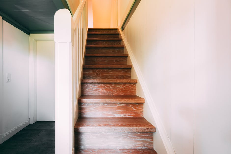 A narrow indoor corridor with white walls and a tiled floor, showing signs of wear and scuff marks. On the left side, there are two small, closed windows with metal frames and a door partially visible